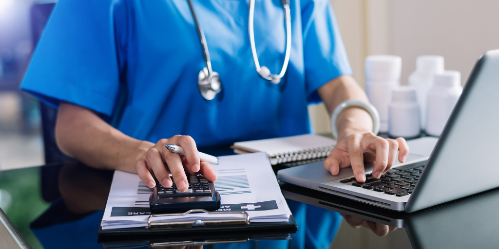 Nurse at a desk using a laptop and calculator with a clipboard 